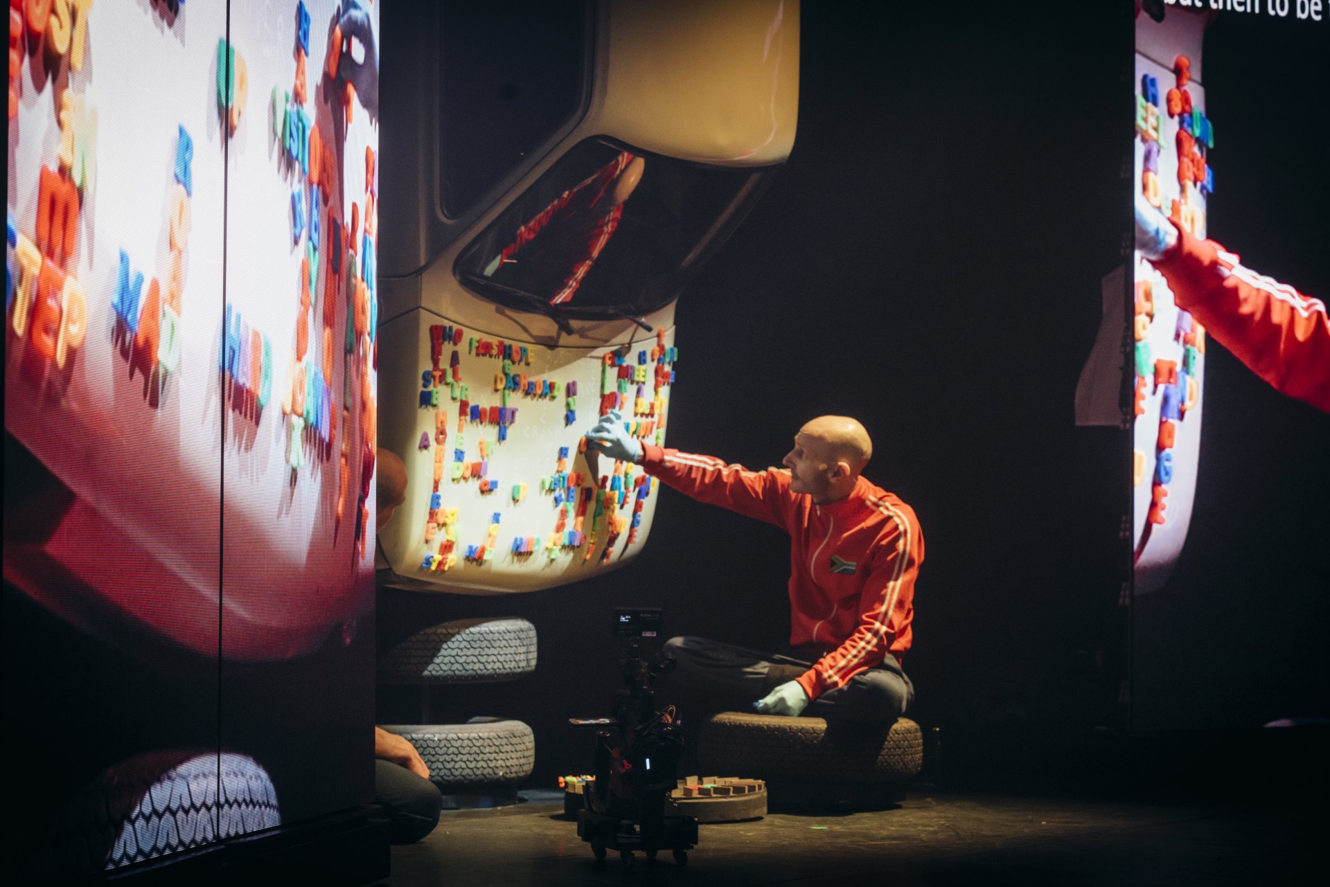 Farbfoto von sitzender Person in roter Trainingsjacke, die magnetische Plastikbuchstaben auf dem Heck eines Autos bewegt. Das Auto hängt senkrecht von der Decke. Zwei Bildschirme rechts und links zeigen einen Zoom auf die Plastikworte (nicht lesbar).