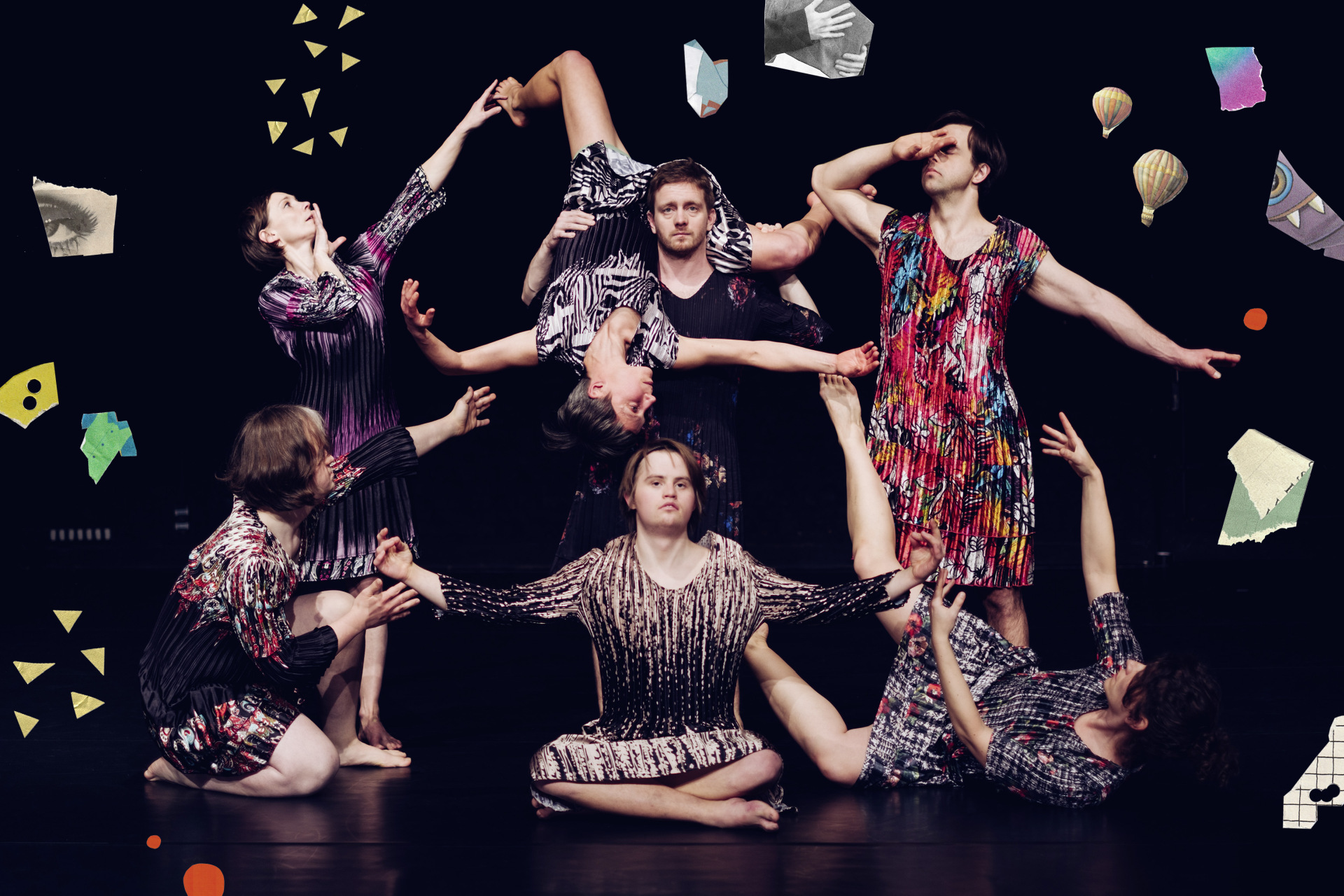 Sieben Tänzer*innen sind lose kreisförmig angeordnet. Das funktioniert, weil sie sich gegenseitig an Armen und Beinen berühren und die vorderen drei Menschen auf dem Boden sitzen. Eine Person wird kopfüber gehalten.