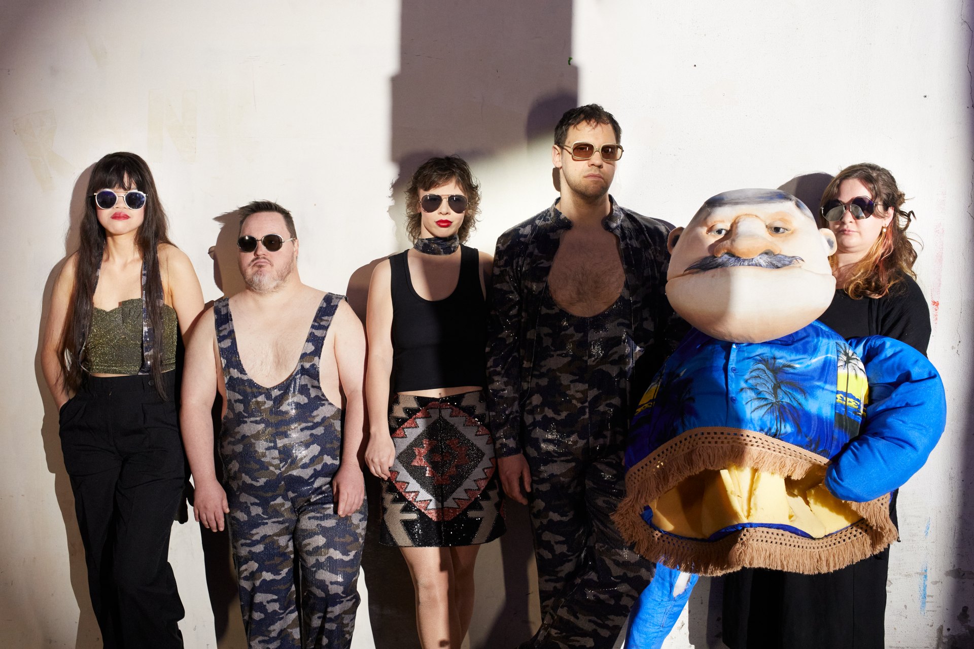 Fünf Personen mit Sonnenbrillen und in rockigen Outfits lehnen an eine Wand. Eine von ihnen hält eine Puppe mit strahlend blauer Jacke und grauem Schnurrbart.
