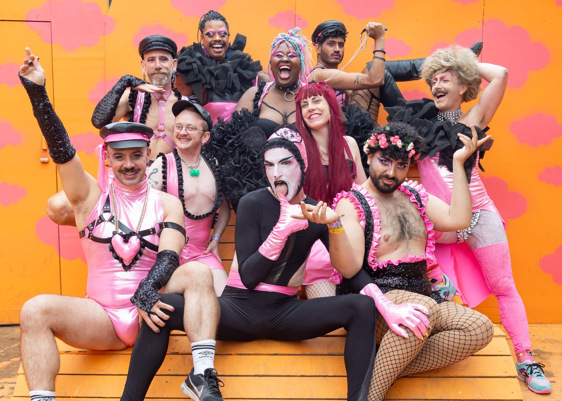 Farbfoto von zehn Drag Performer*innen. Alle tragen sexy Kleidung aus rosa Satin, mit schwarzen Rüschen, Leder-Harnessen und Netzstrümpfen. Die meisten lachen in die Kamera, die Person vorne in der Mitte leckt lasziv über ihren Daumen.