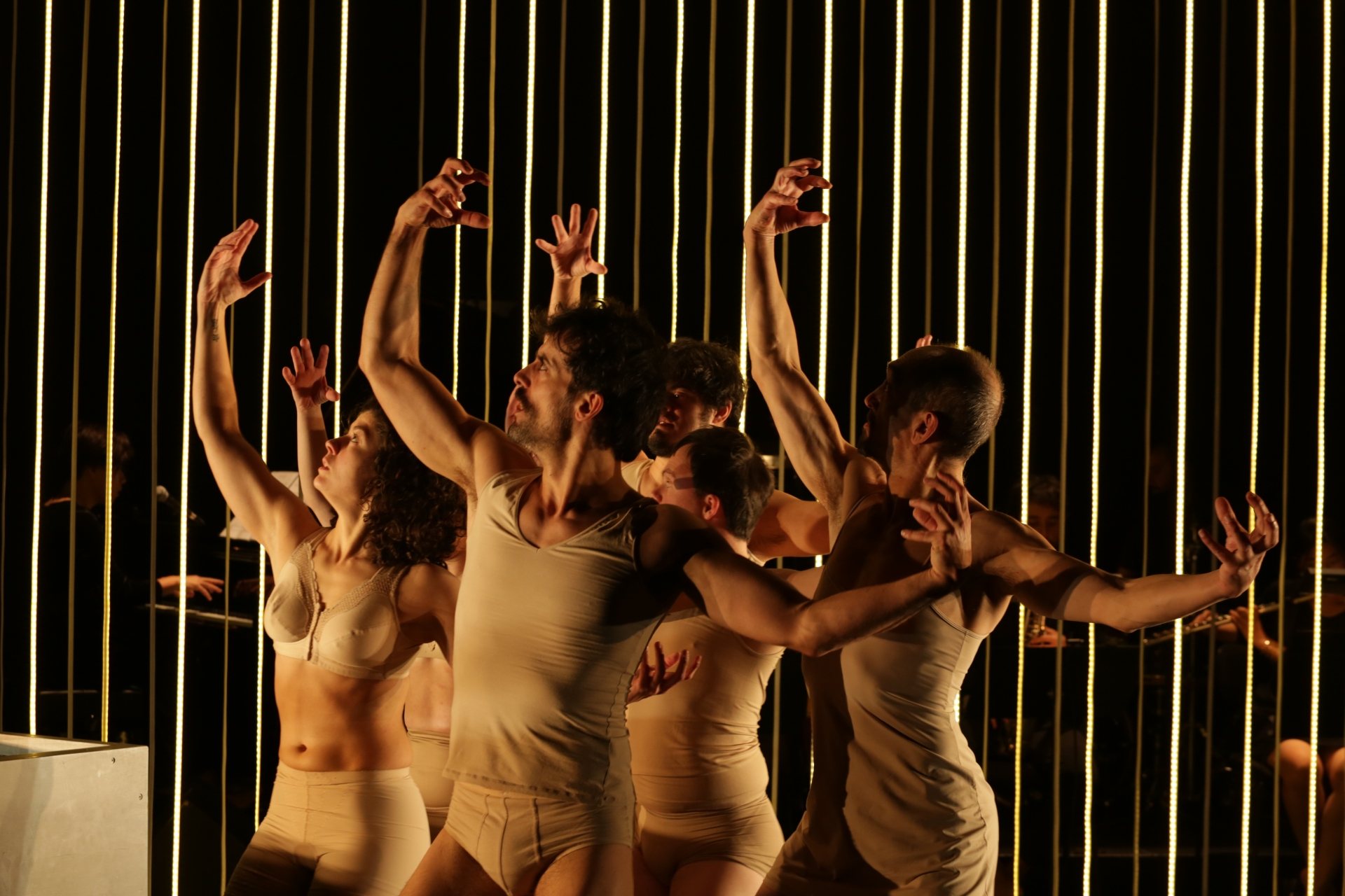 Sechs Personen vor einem Hintergrund aus vertikalen LED Schnüren. Die sechs befinden sich in der gleichen Pose, alle tragen beige Unterwäsche, rechten Arm über den Kopf gehoben, den Linken zur Seite gestreckt.