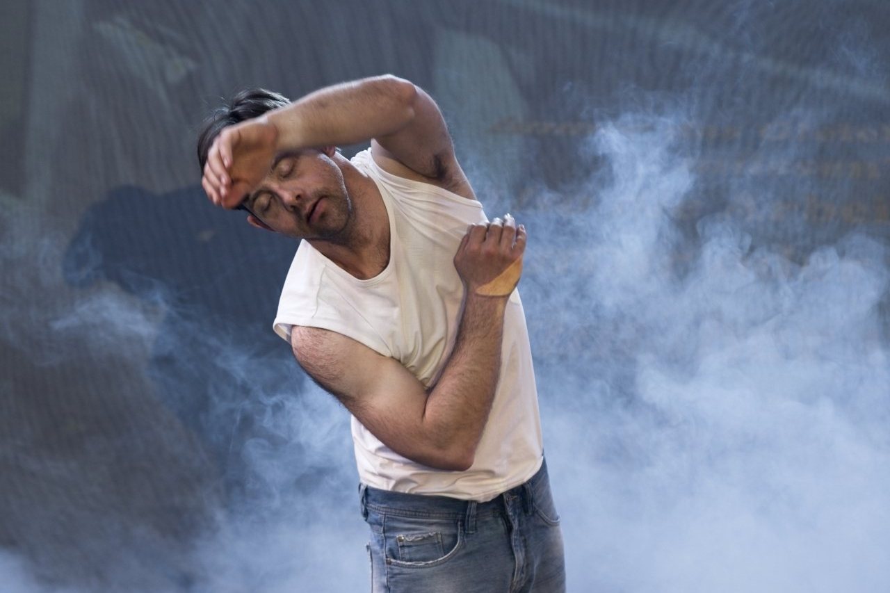 Der Tänzer steht in Kunstnebel, den linken Arm vor den Kopf gestreckt, den rechten an die Brust gelegt. Er trägt Jeans-Shorts und ein weißes Muscle-Shirt.