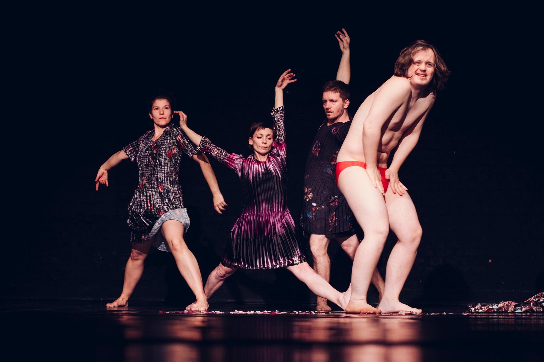 Vier tanzende Personen, die drei Personen links tragen schillernde Kleider, die Person vorne rechts eine rote Unterhose.