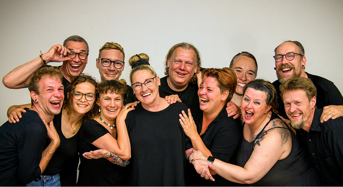 Gruppenfoto von 12 Menschen in schwarzen T-Shirts, die in die Kamera lachen. Der Hintergrund ist weiß.