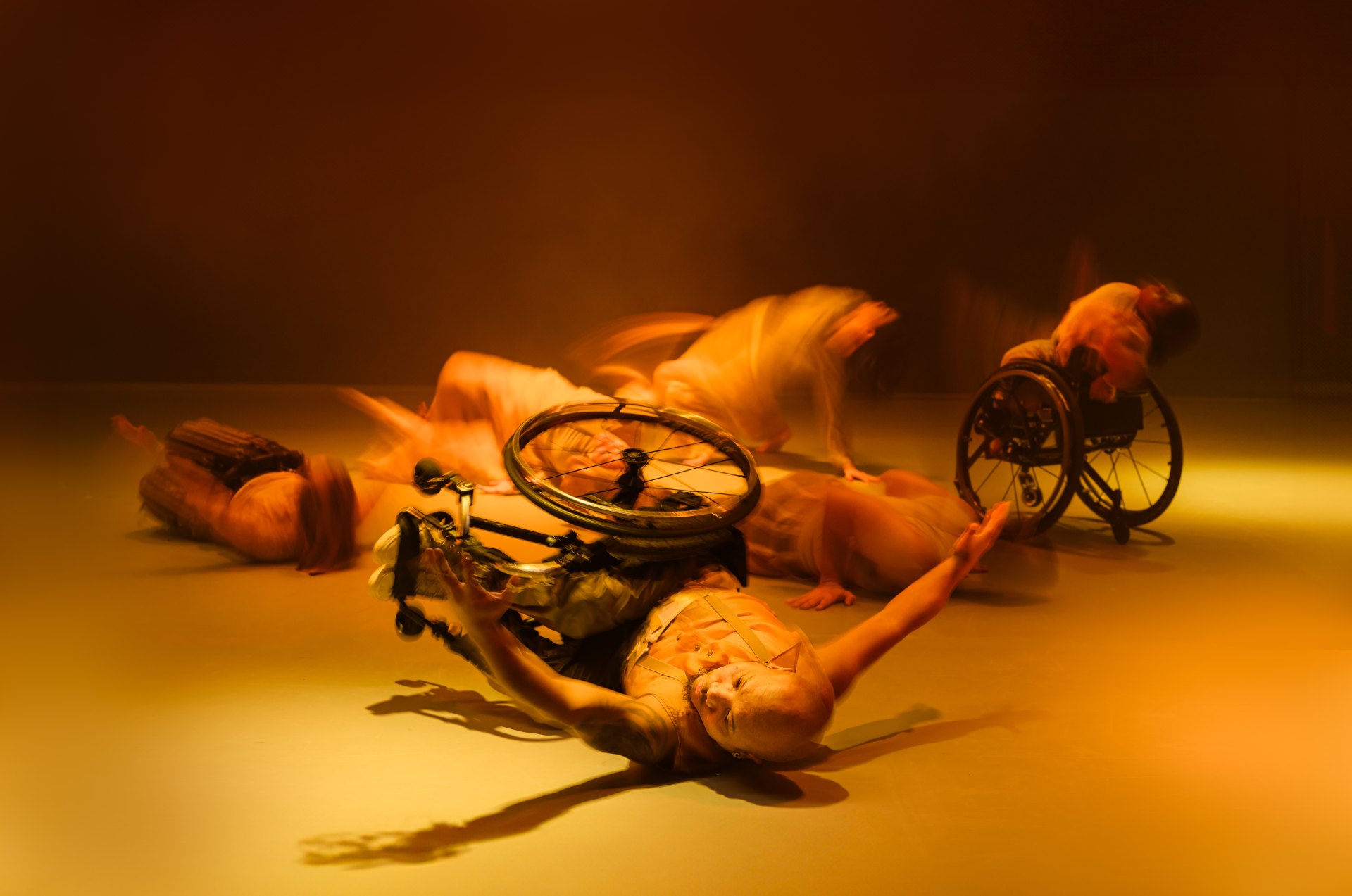 Das Foto ist in gelb-orangenem Licht ausgeleuchtet. Darauf sind sechs Tänzer*innen zu sehen, die auf dem Boden liegen oder im Rollstuhl sitzend sich rückwärts ins Hohlkreuz beugen.